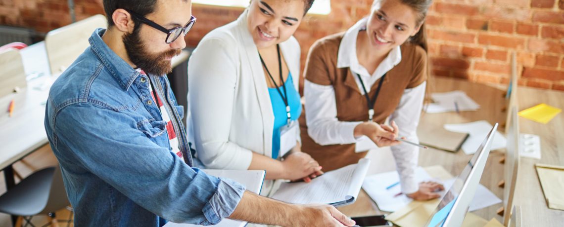Young bearded confident analyst pointing at computer screen while making presentation of financial data to colleagues